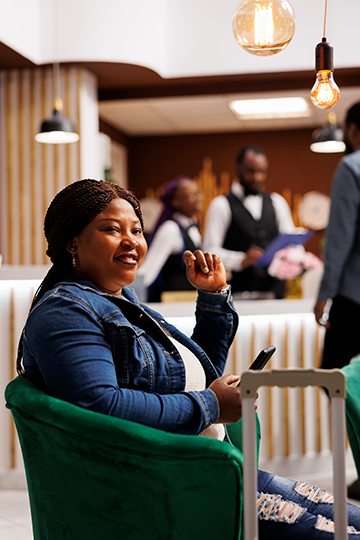 happy-smiling-african-american-woman-arriving-resort-sitting-hotel-lobby-with-smartphone-waiting-until-check-time-black-female-guest-sitting-lounge-area-using-phone-traveling-alone